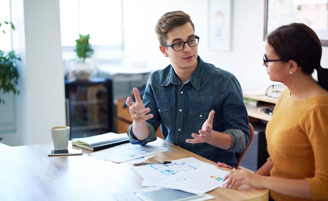 Two people sitting at a table with papers on top in an office talking to each other.