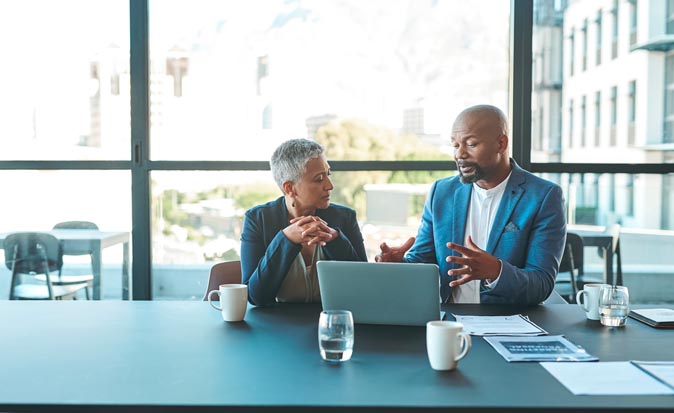 Two corporate executives talking at a conference table with a laptop computer in front of them.