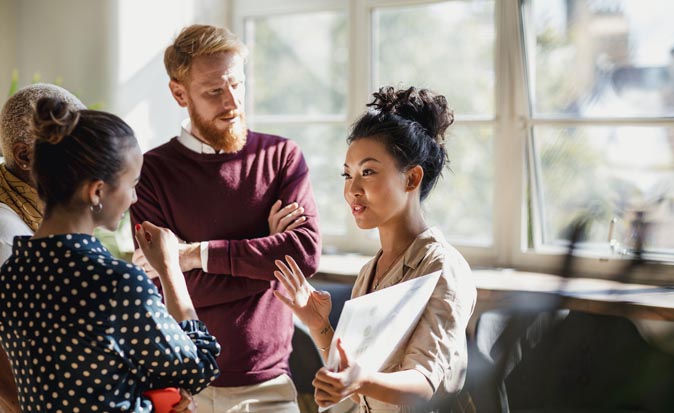 Four people talking in an office. One is holding documents and gesturing with hands as others listen.