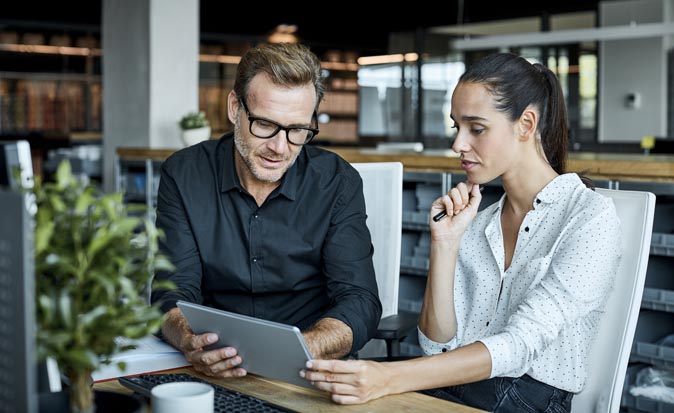 Two business leaders sitting at a desk in an office looking at a tablet computer they are holding.