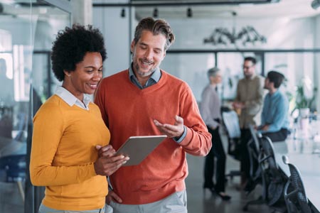 Two managers looking at a tablet computer in an office while several others talk in background.
