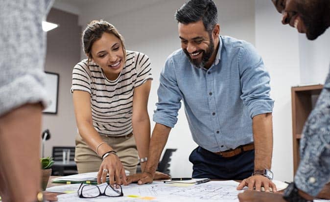 A group of managers smiling at blueprints on table in an office during a strategic planning meeting.