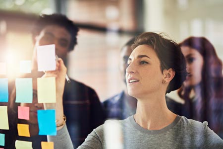 A female manager writing on a sticky note in front of a glass wall while a group of people stand behind her.