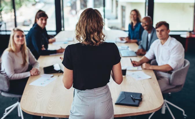 A woman standing in front of a group of people around a table practicing her presentation skills.