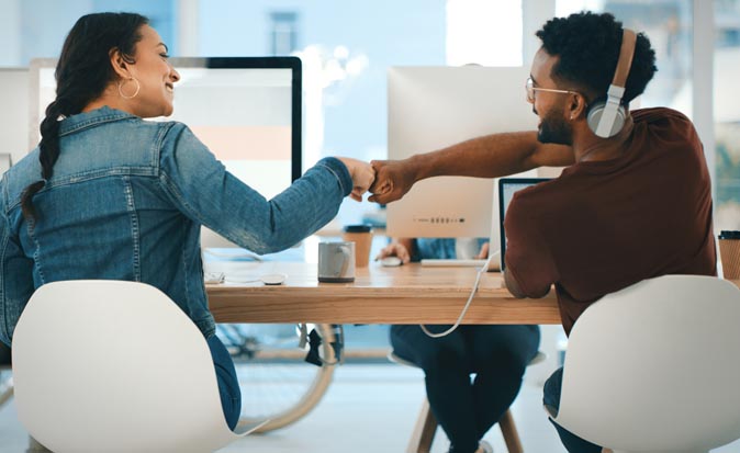 Two co-workers sitting next to each other reaching out their arms to fist bump to celebrate.