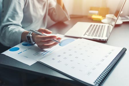 A woman sitting at a desk has good time management by writing on a calendar while using a laptop.